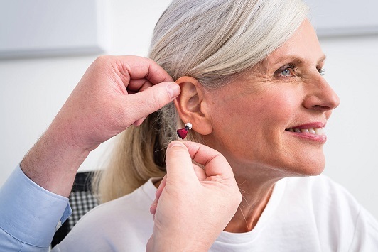 Ältere Frau bekommt ein modernes Hörgerät ins Ohr eingesetzt und lächelt dabei.